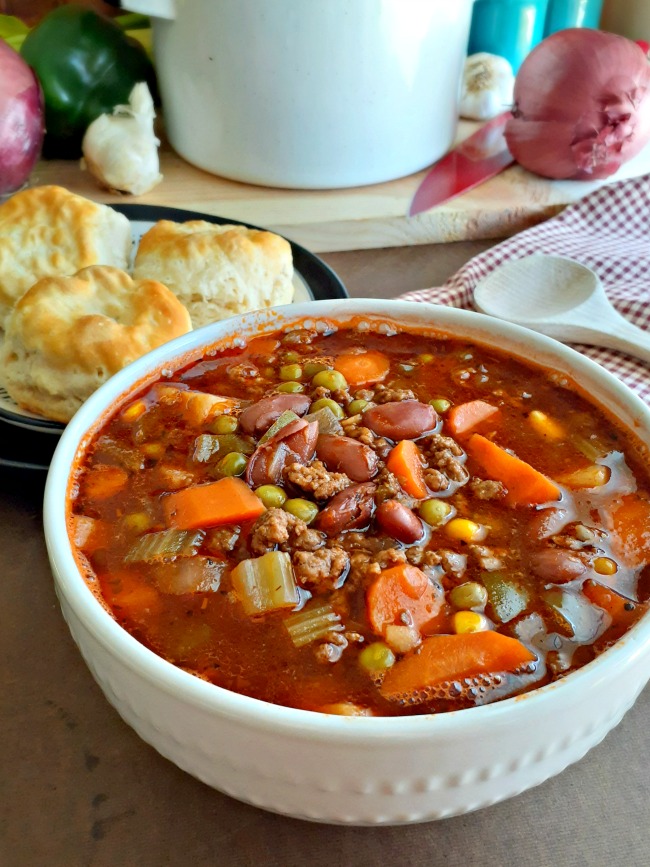 This photo shows Country Beef Vegetable Soup served in a white bowl with a plate of biscuits next to the bowl.