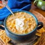 Creamy corn soup in a teal bowl topped with sour cream and shredded cheese, served with crackers on a wooden board.