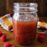 Glass jar filled with raspberry jam on a wooden table, with fresh raspberries nearby and a colorful cloth in the background.