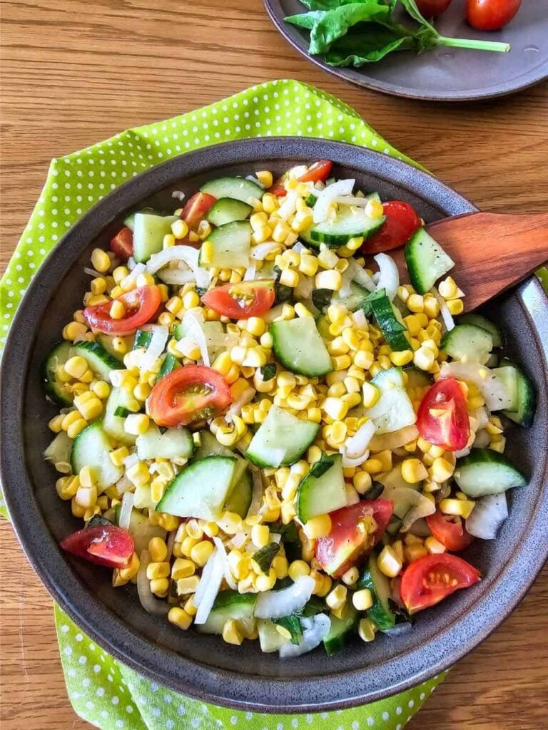 Bowl of fresh corn, cucumber, tomato, and onion salad on a gray plate with a wooden spoon.