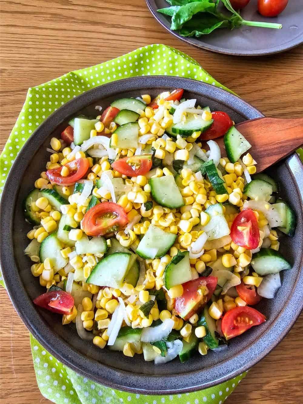 Bowl of fresh corn, cucumber, tomato, and onion salad on a gray plate with a wooden spoon.